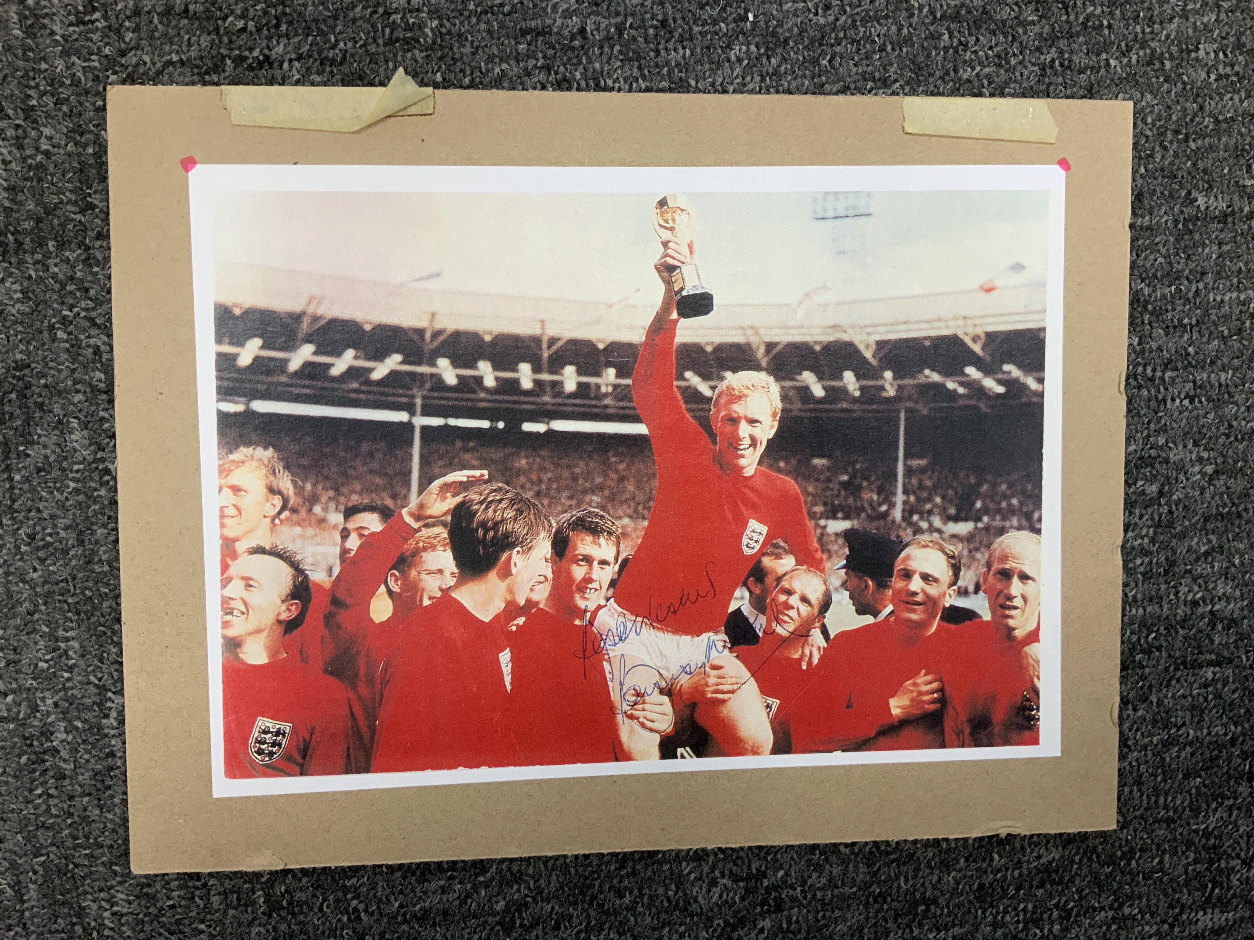 Bobby Moore Signed England Football Print: An A4 print of the famous picture of Moore being held aloft holding the World Cup. Hand signed in blue biro Best Wishes with no dedication. Mounted on board.
