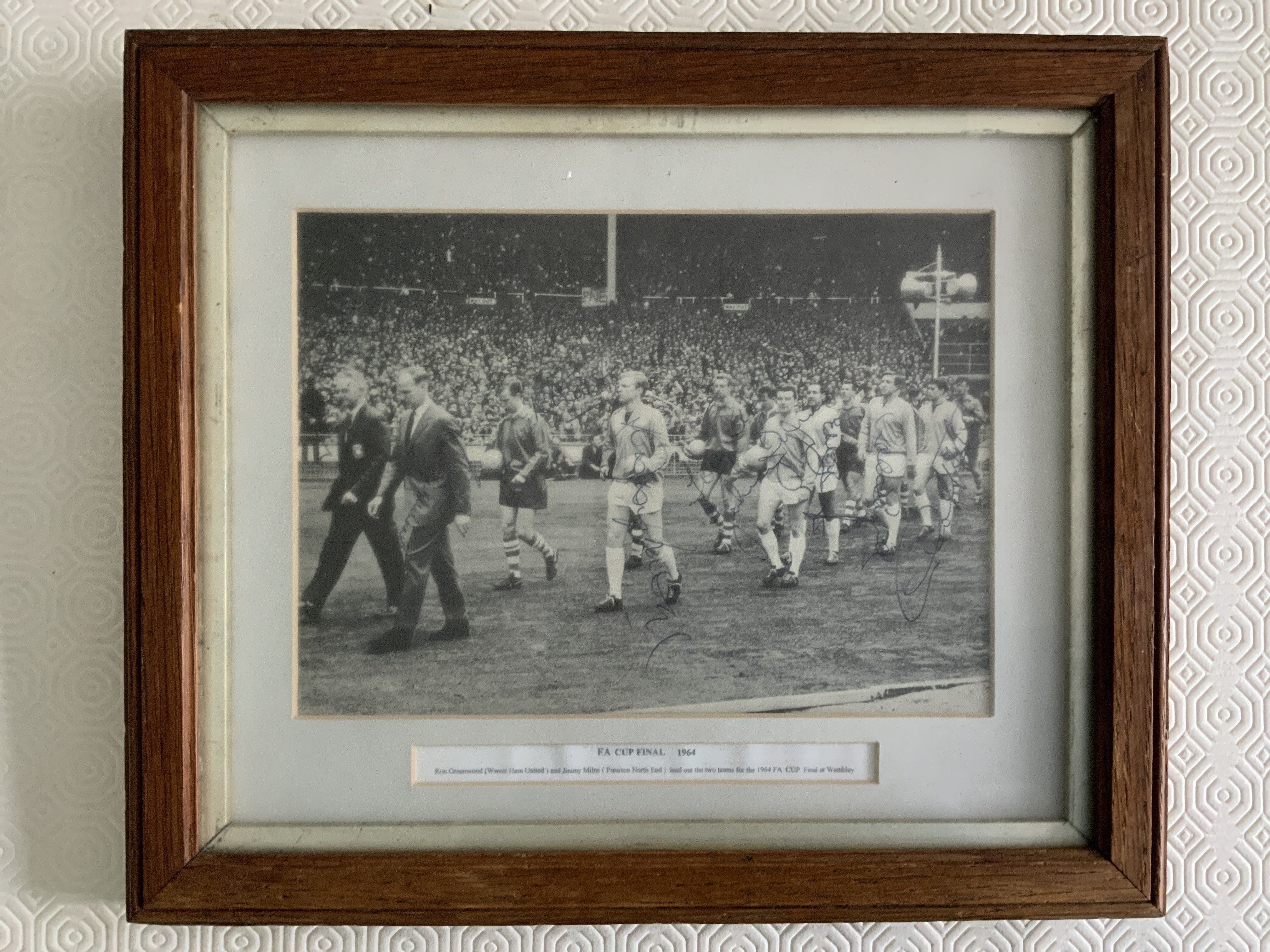 1964 West Ham Signed Framed Cup Final Picture: Great shot of Greenwood leading the teams out at Wembley with all 5 West Ham players having signed. Autographs of Greenwood, Moore, Byrne, Bovington, Bond and Hurst. Mounted and framed many years ago with genuine signatures. Measures just over 13 x 11 inches.